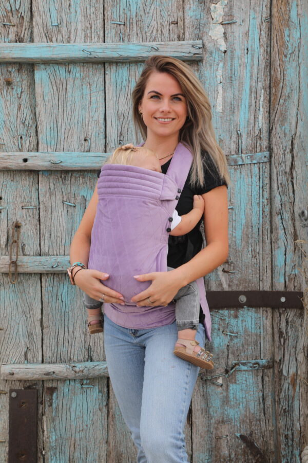 Mother carries babygirl in lilac velvet baby carrier with checkered motif