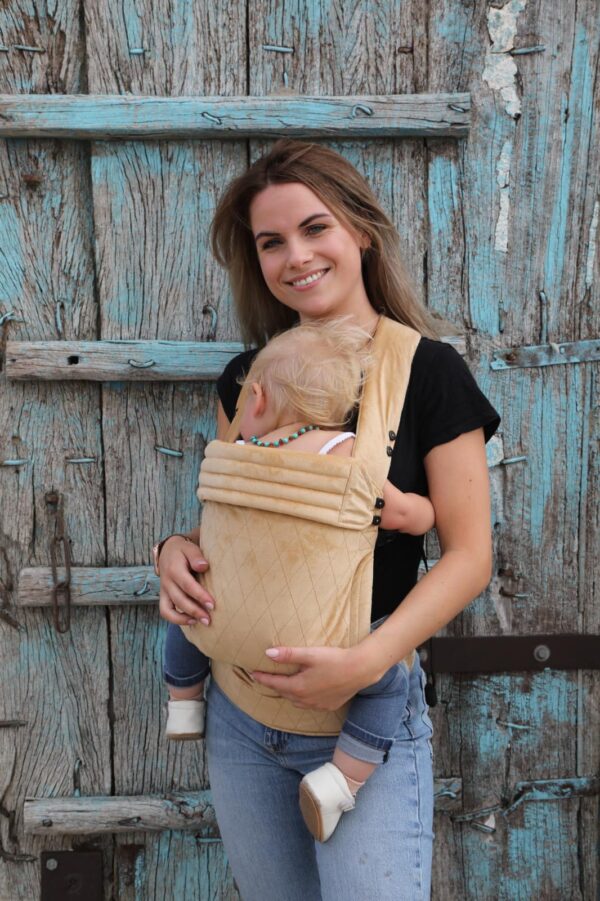 Mother carries her baby daughter in a velvet beige baby carrier with checkered motif in front of a porch